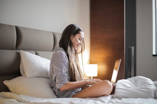 Foto De Vista Lateral De Uma Mulher Sorridente Em Uma Blusa Listrada De Preto E Branco, Sentada Em Uma Cama Enquanto Usa Um Laptop