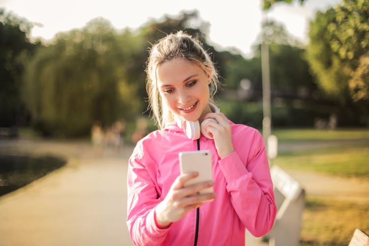 Selective Focus Photo Of Smiling Woman In A Pink Jacket With White Headphones On Her Neck Using A White Phone 