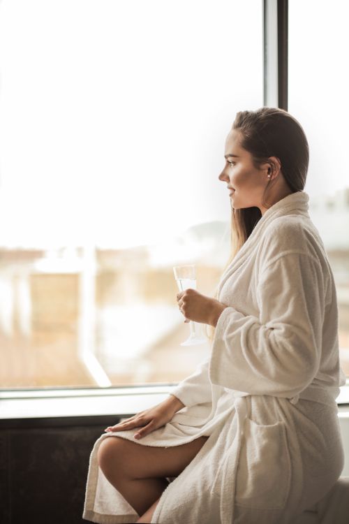 Free A serene woman in a bathrobe gazes out of a window while holding a champagne glass. Stock Photo