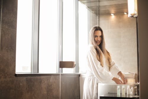 Photo of Woman in White Bathrobe Standing in the Bathroom While Washing Her Hand at a Sink