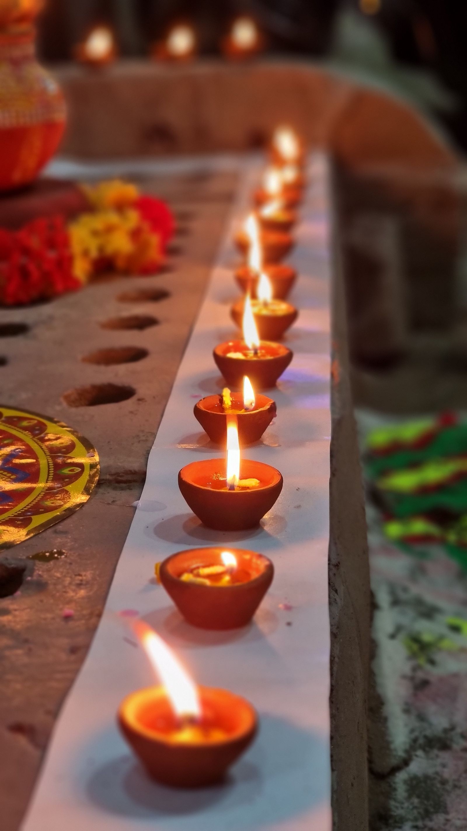Clay oil lamps burning in a line beside marigold garlands and a painted rangoli on a ledge at dusk