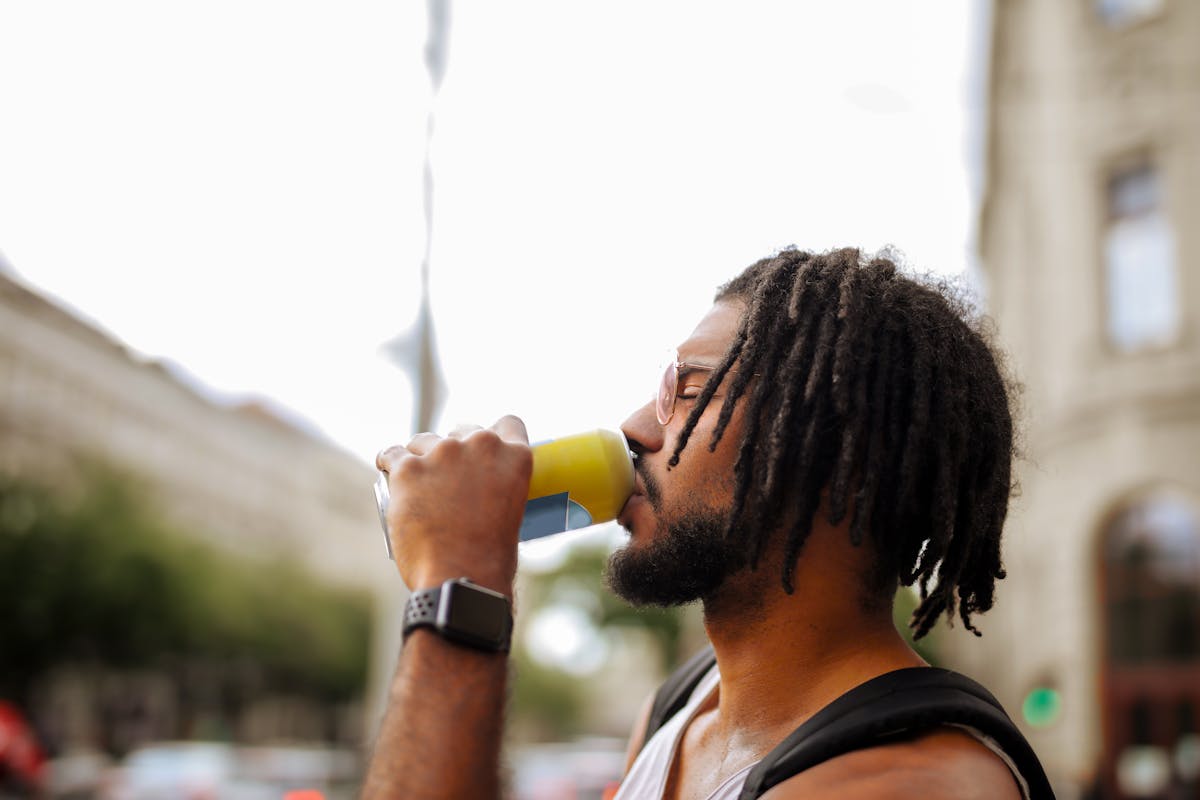 Side view of adult Hispanic guy with dreadlocks in sunglasses and casual clothes with backpack and smart watch drinking yummy beverage from vivid yellow can while standing with eyes closed on street in downtown