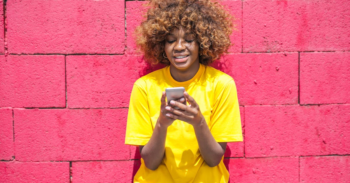 Young woman with curly hair using a smartphone against a bright pink brick wall, smiling outdoors.