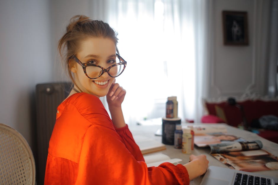 Joyful female in glasses and vivid orange clothes smiling at camera while sitting at table with laptop and magazines against blurred interior of light modern living room