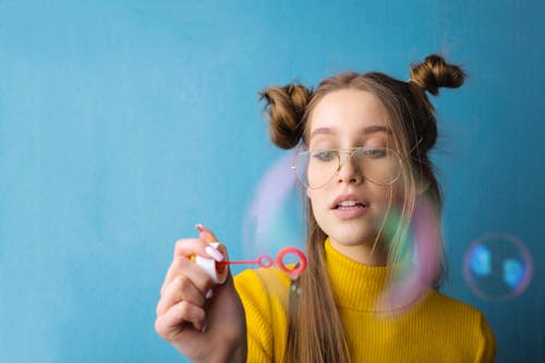 Foto Ritratto Di Donna In Maglione Dolcevita Giallo E Occhiali Da Vista Che Soffia Bolle Stando In Piedi Davanti A Sfondo Blu