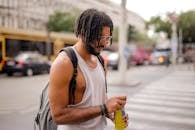 Confident adult Hispanic male traveler in sunglasses and casual clothes with backpack opening can of drink while walking along pavement against blurred cars driving along street and empty crosswalk in downtown