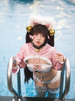 A young woman in a pink cat-themed cosplay outfit poses by a swimming pool ladder.