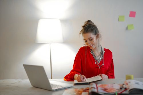 Free Smiling woman in red top writing in notebook at desk with laptop and lamp indoors. Stock Photo