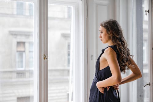 Photo of Woman in Black Dress Standing Beside Glass Window looking Outside