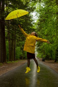 Woman in yellow raincoat and boots jumps joyfully with umbrella on a wet forest path.