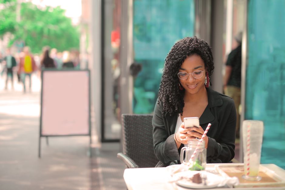 Photo by Andrea Piacquadio on Pexels A woman sits outdoors at a café, smiling while looking at her smartphone.