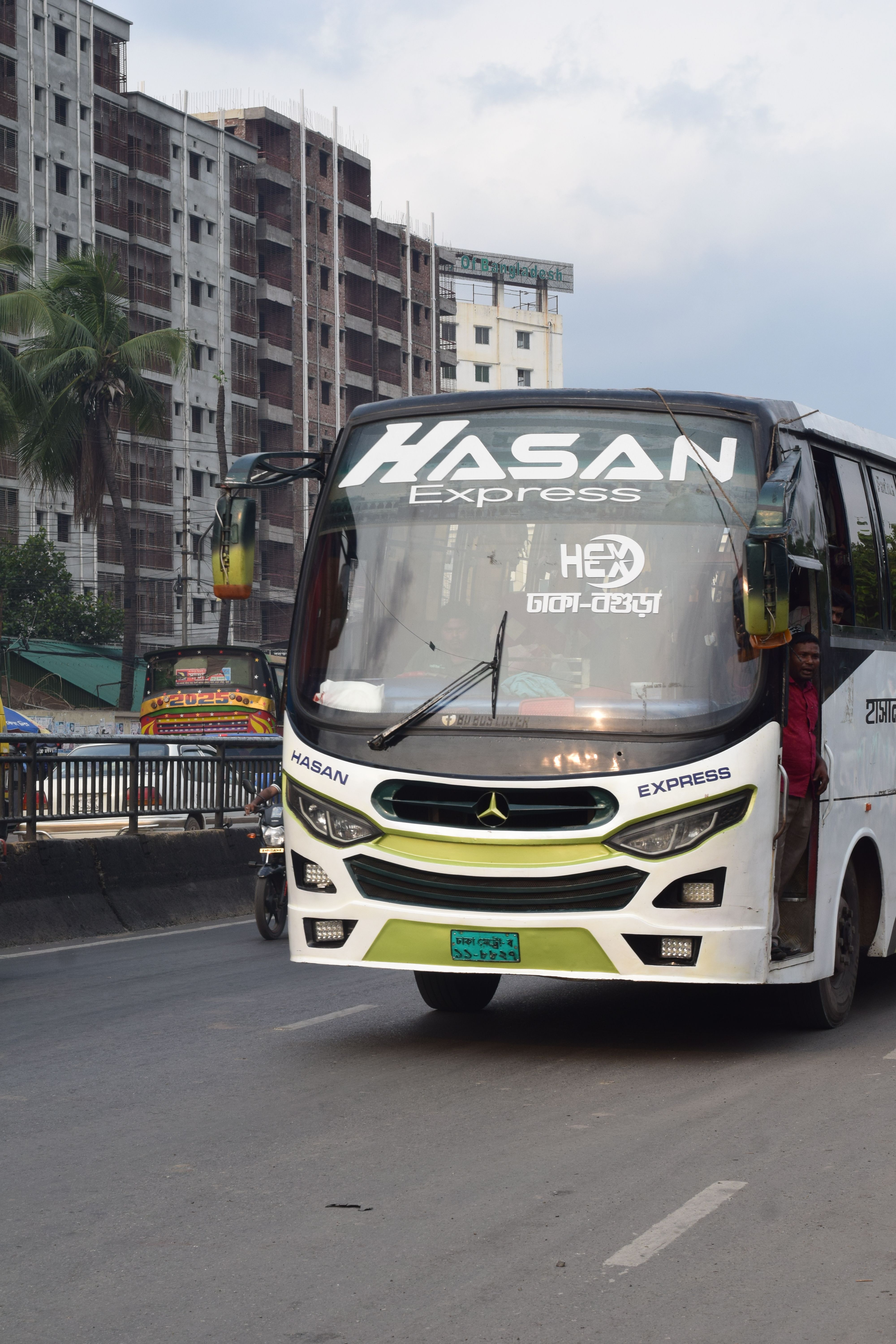 A Hasan Express bus driving through Aminbazar, Dhaka, capturing urban transit in Bangladesh.