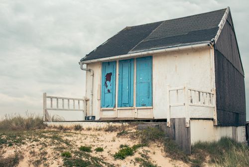 Free A quaint, weathered beach hut with blue doors on a sandy dune under a cloudy sky in Hauts-de-France. Stock Photo