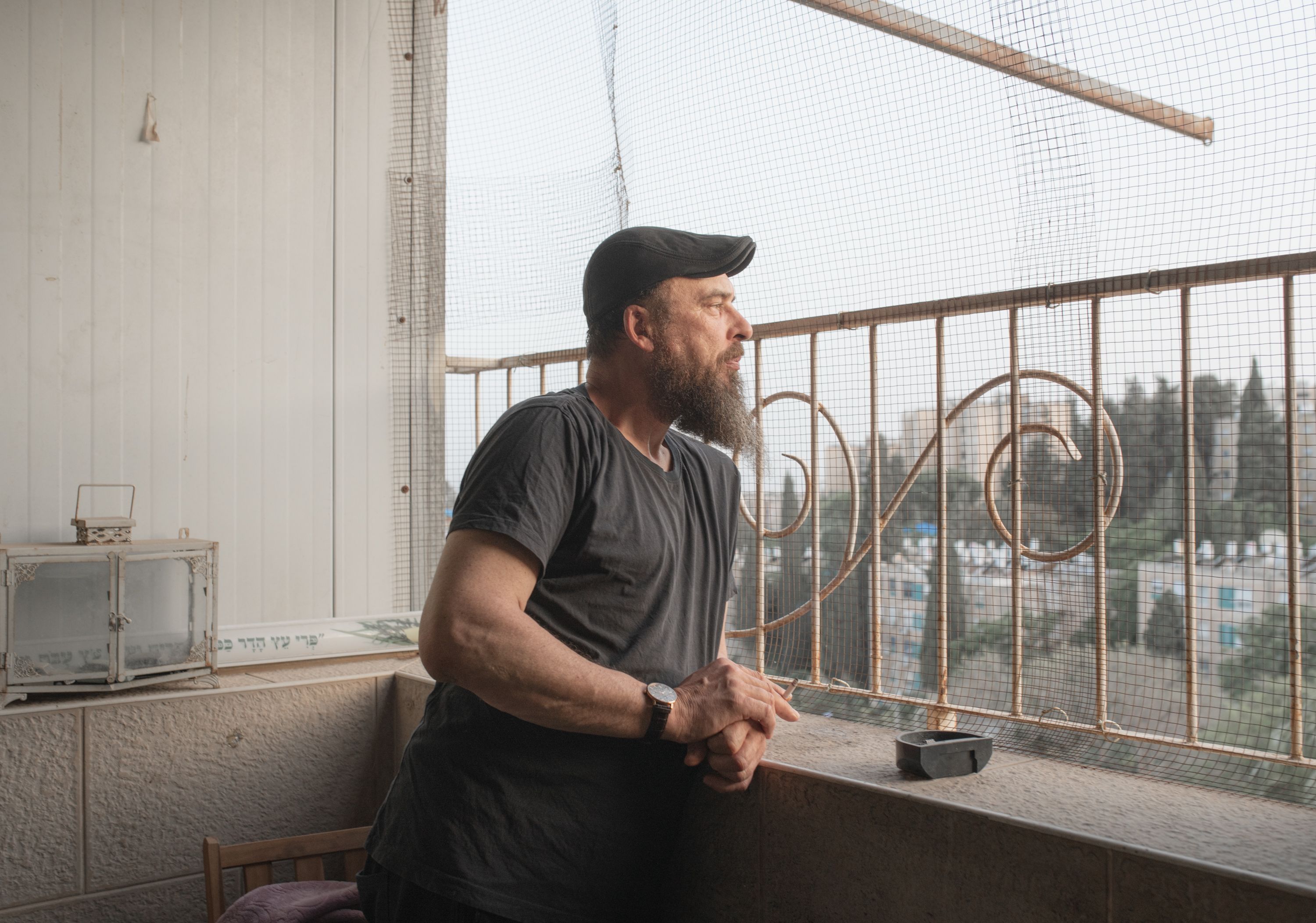 A man in a black cap stands pensively on a balcony during daytime.