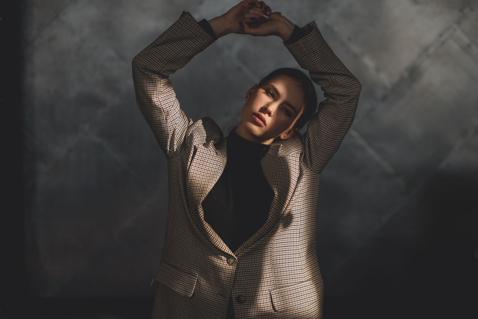 Woman in a checked blazer with arms raised overhead, warm sunlight falling across her face