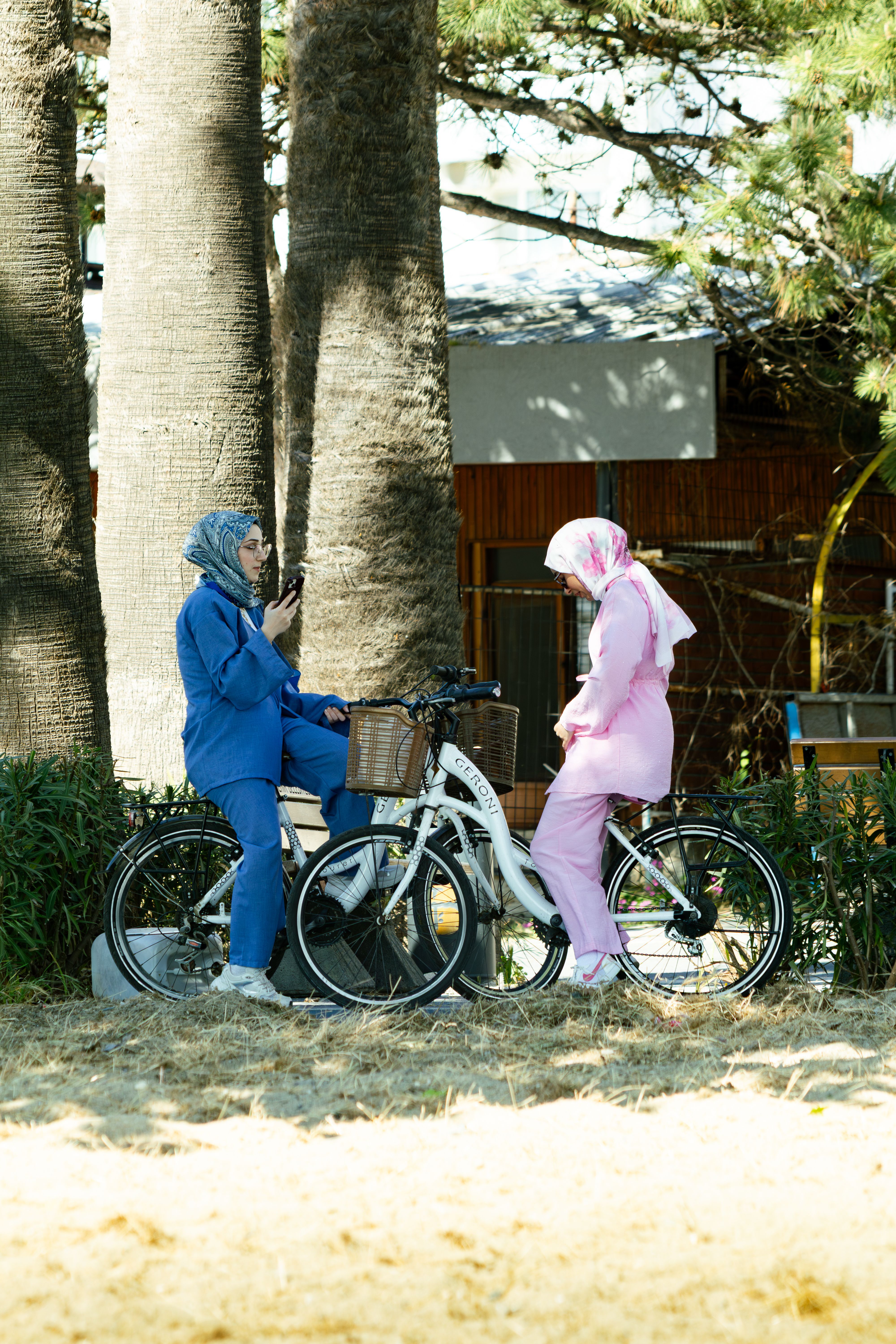 Two women in colorful outfits with bicycles conversing among trees in a park.