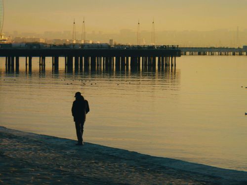 Free A person walks by a serene beach with a dock in the background at sunset, creating a peaceful scene. Stock Photo