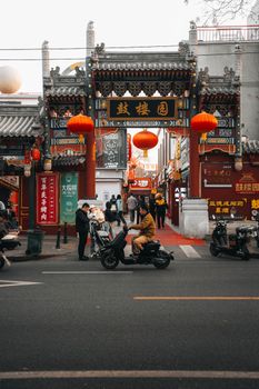 Bustling street scene at a traditional Chinese market entrance with red lanterns and people passing by.