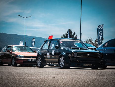 A line-up of vintage and modern cars participating in a race event under clear skies with mountains in the background.
