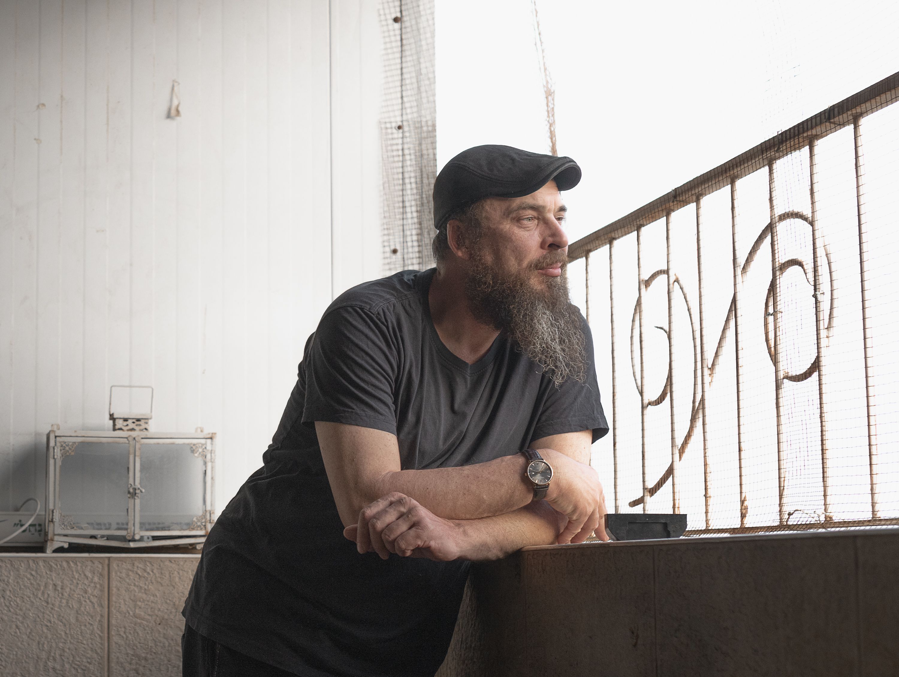 A bearded man wearing a black hat and shirt leans on a balcony railing, gazing thoughtfully.