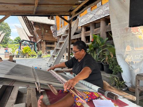 Indonesian woman weaving under traditional wooden houses, showcasing local craftsmanship and cultural heritage.