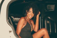 一位卷发的时尚年轻女子坐在车内，微笑着展示别致的服装