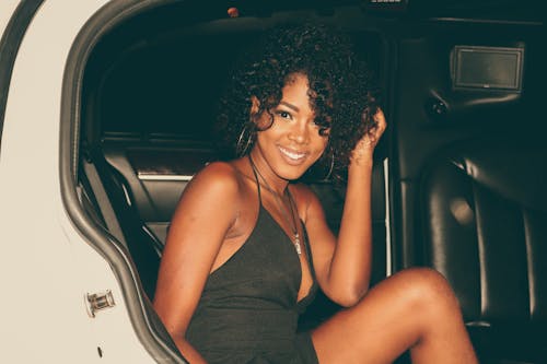 A fashionable young woman with curly hair sits smiling inside a car, showcasing a chic outfit.
