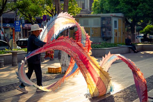 A senior man skillfully handles a colorful dragon kite in a sunlit urban park.