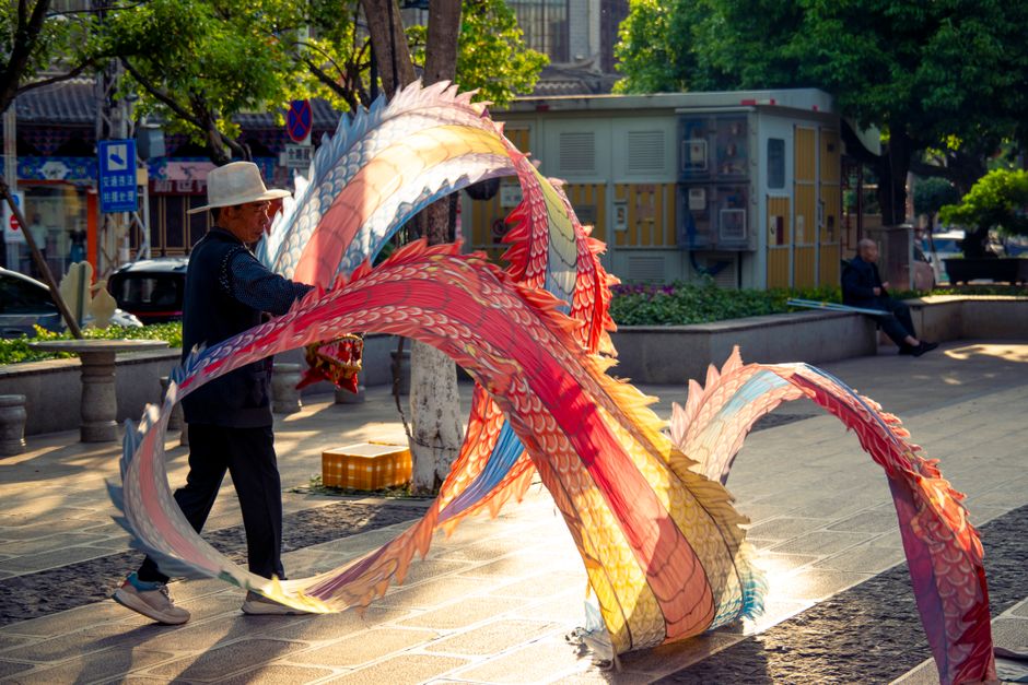 A senior man skillfully handles a colorful dragon kite in a sunlit urban park.