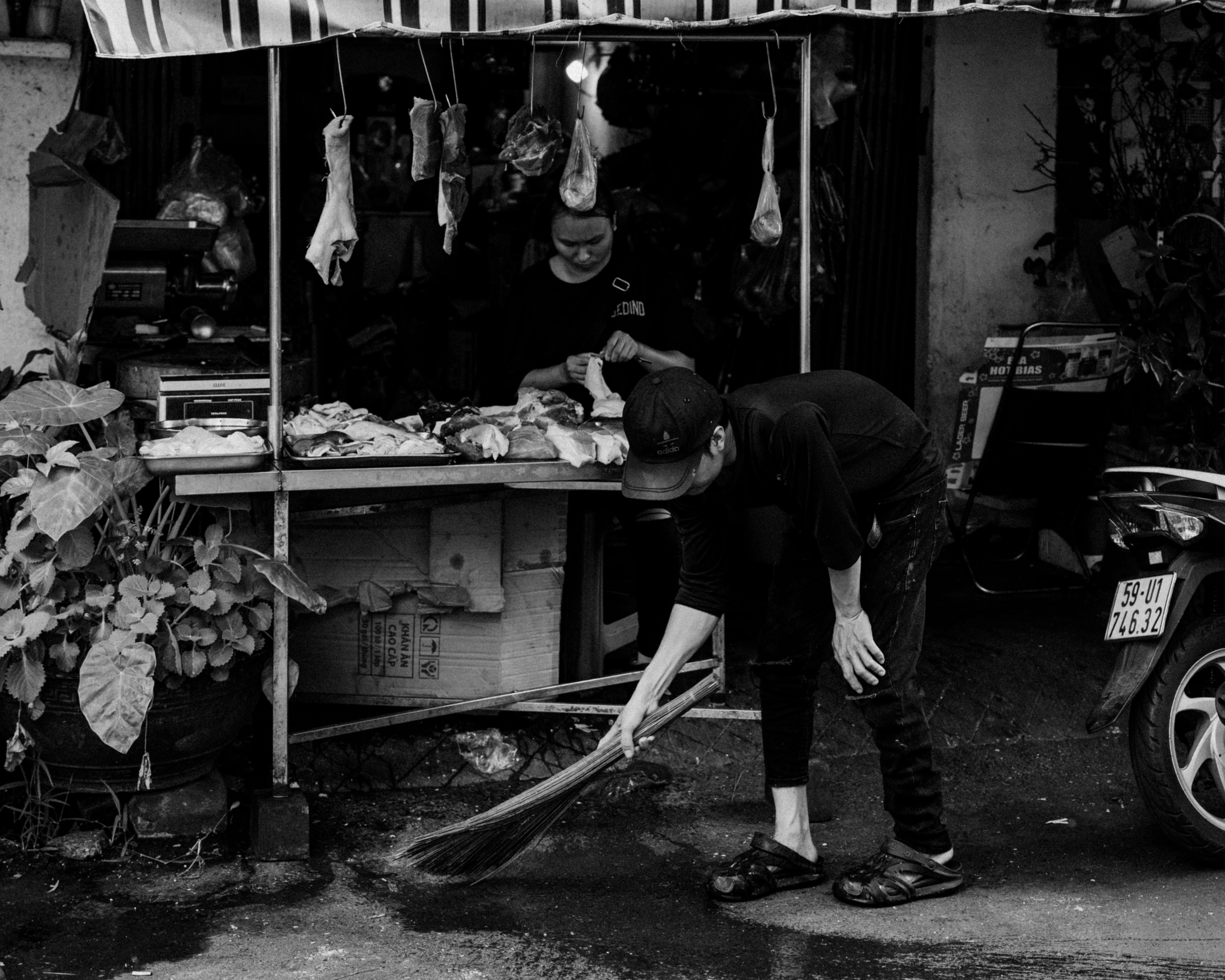 Black and white photo of a market vendor cleaning outside a shop.