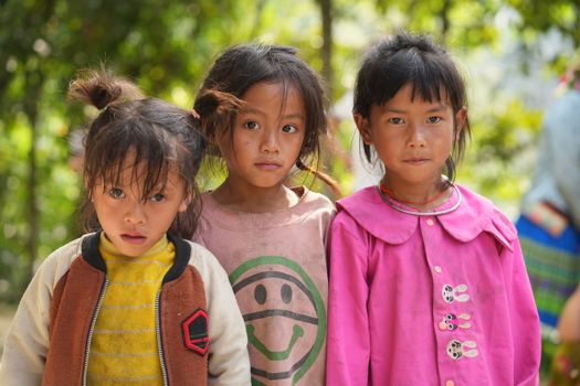 Three young children stand in colorful clothes against a lush outdoor backdrop.