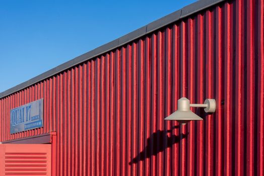 Close-up of a red warehouse wall featuring a light fixture against a clear blue sky.
