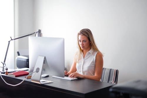 Woman in White Sleeveless Shirt Using Macbook Air on Brown Wooden Table