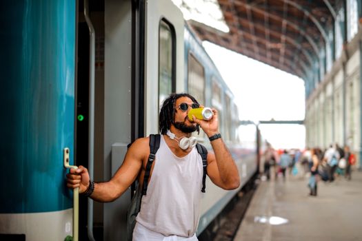 A young man with headphones drinks a beverage while standing on a train platform.