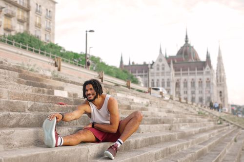 Free Man with dreadlocks smiling while stretching on city steps near architectural landmark. Stock Photo