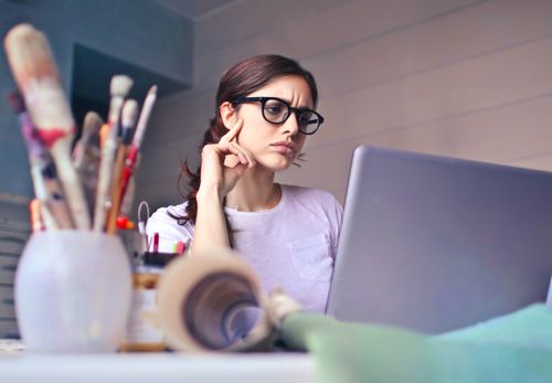 Free Focused woman with glasses working on laptop in an artistic workspace with paintbrushes. Stock Photo