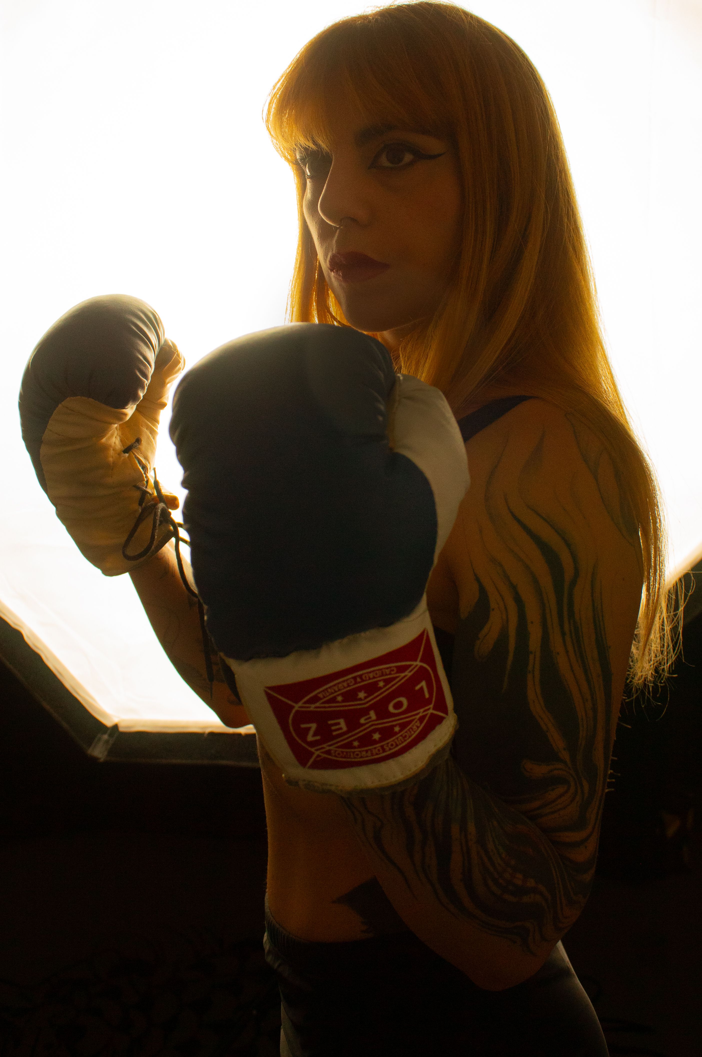 A strong and determined female boxer with tattoos poses with boxing gloves under dramatic lighting.