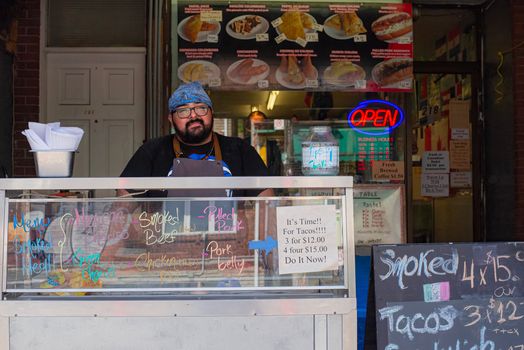 Man at taco stand with neon open sign, various menu items, and promotional offers.