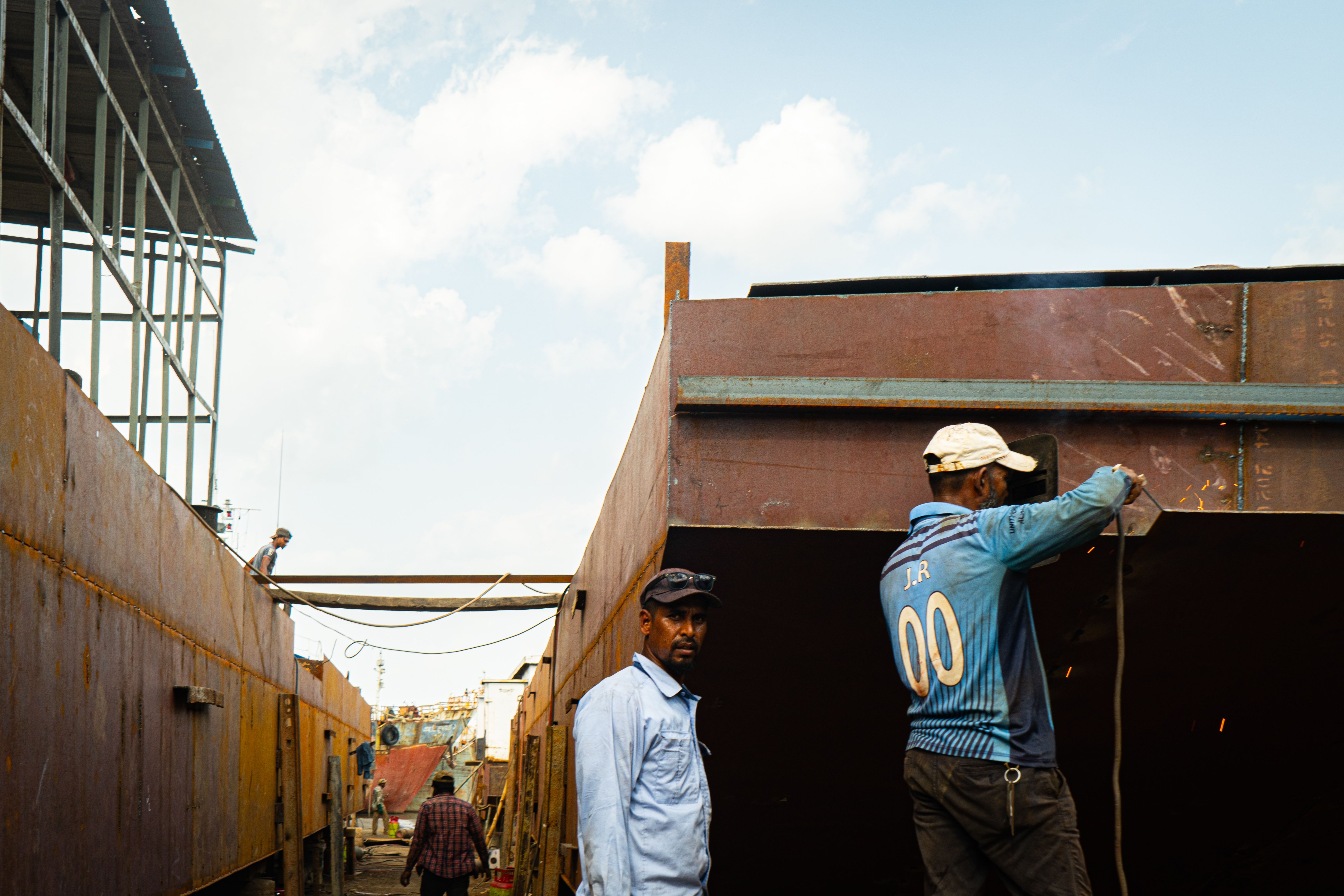 Two shipyard workers performing welding work on a vessel under bright daylight.