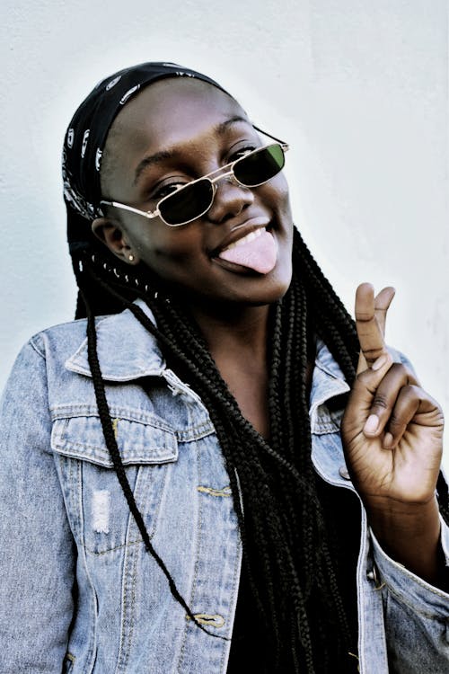 Photo of Woman in Blue Denim Jacket Wearing Black Sunglasses Posing with Her Tongue Out While Doing the Peace Sign