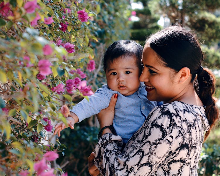 Smiling Mother Carry Her Cute Baby Boy Near The Blooming Flowers