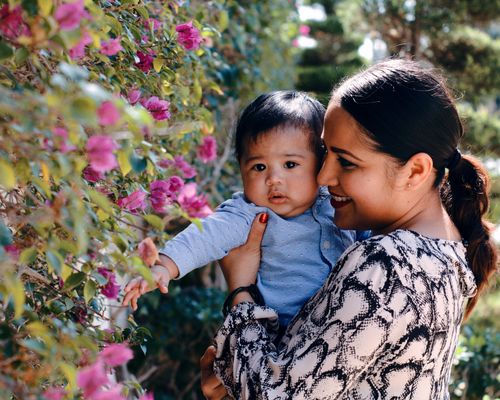 Free Cheerful young ethnic female hugging cute baby boy and smiling while spending time together in garden near flowers Stock Photo
