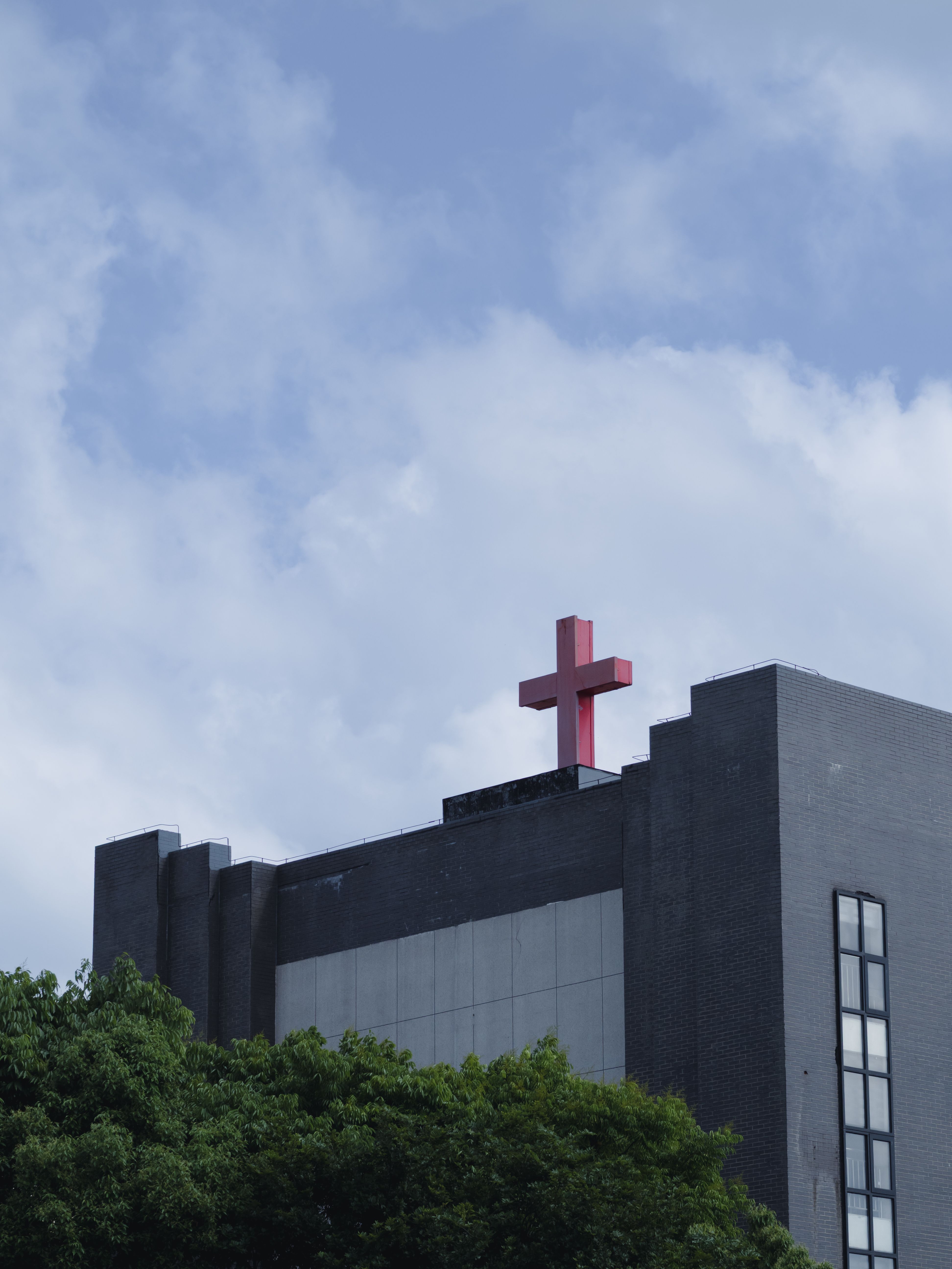A modern gray church building with a bold red cross on its roof, set against a cloudy blue sky.