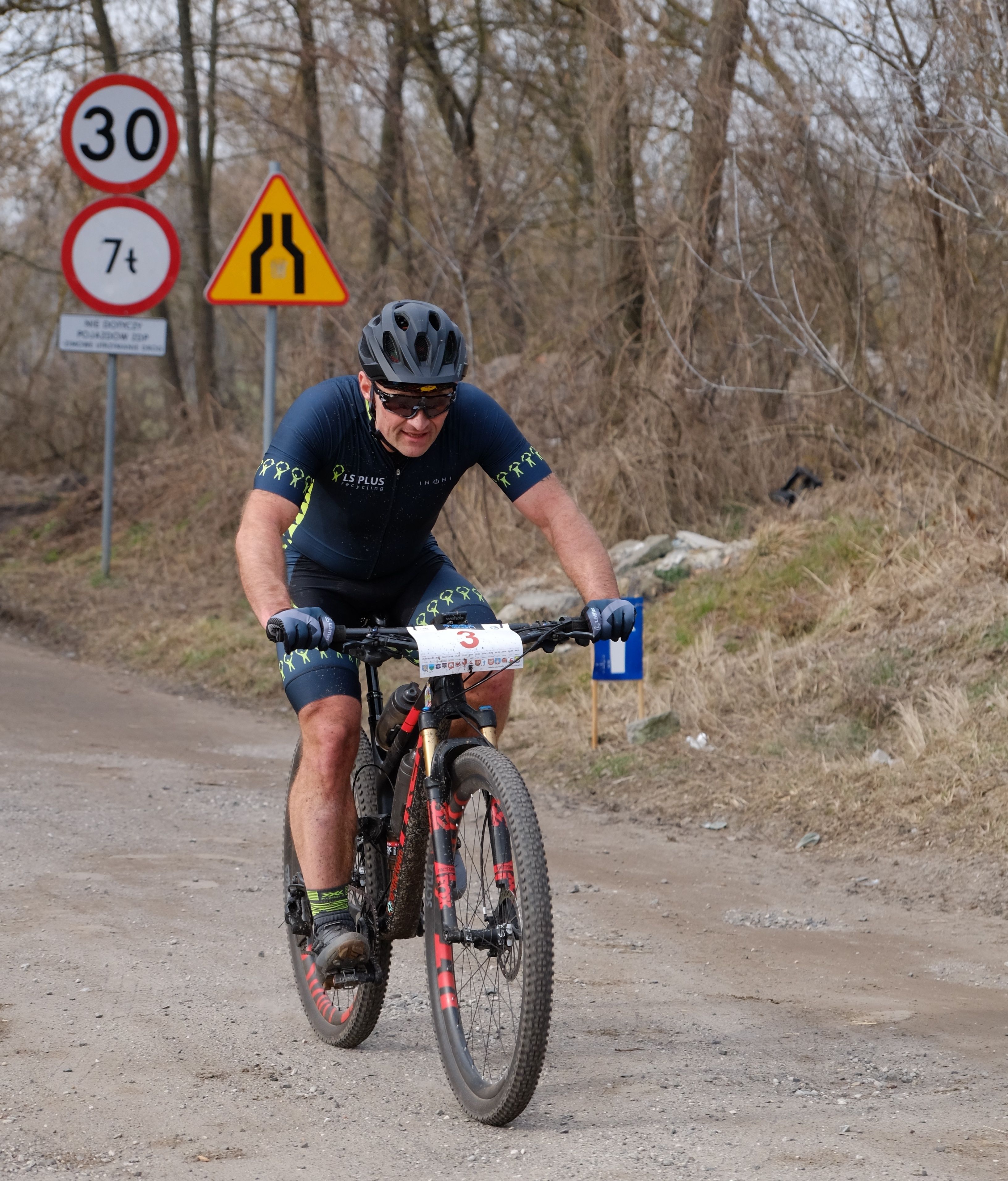 Cyclist participates in a rural bike race, wearing protective gear. Road sign indicates 30 km/h limit.