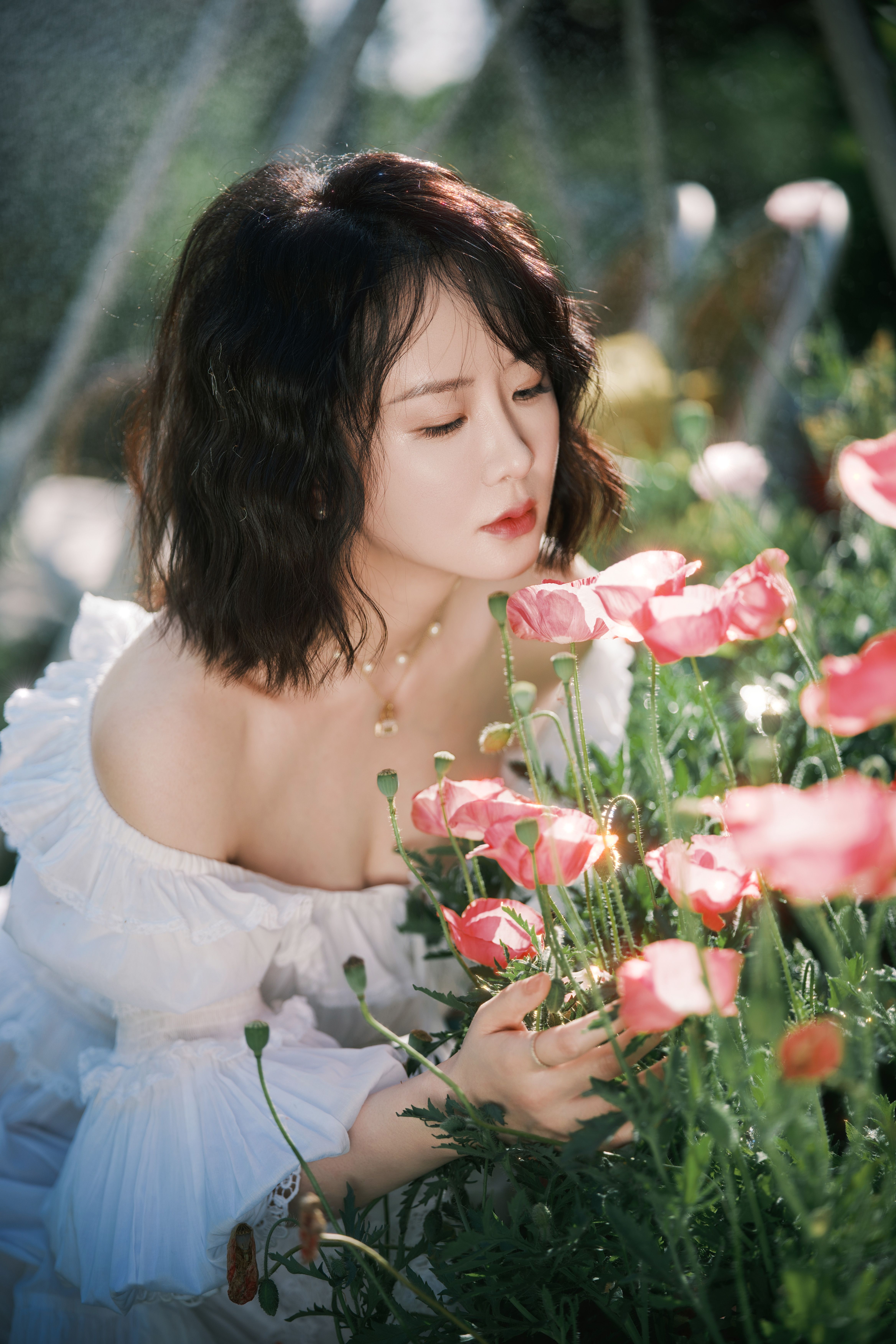 A young woman in a white dress admires vibrant pink flowers in a sunlit garden, enjoying nature.