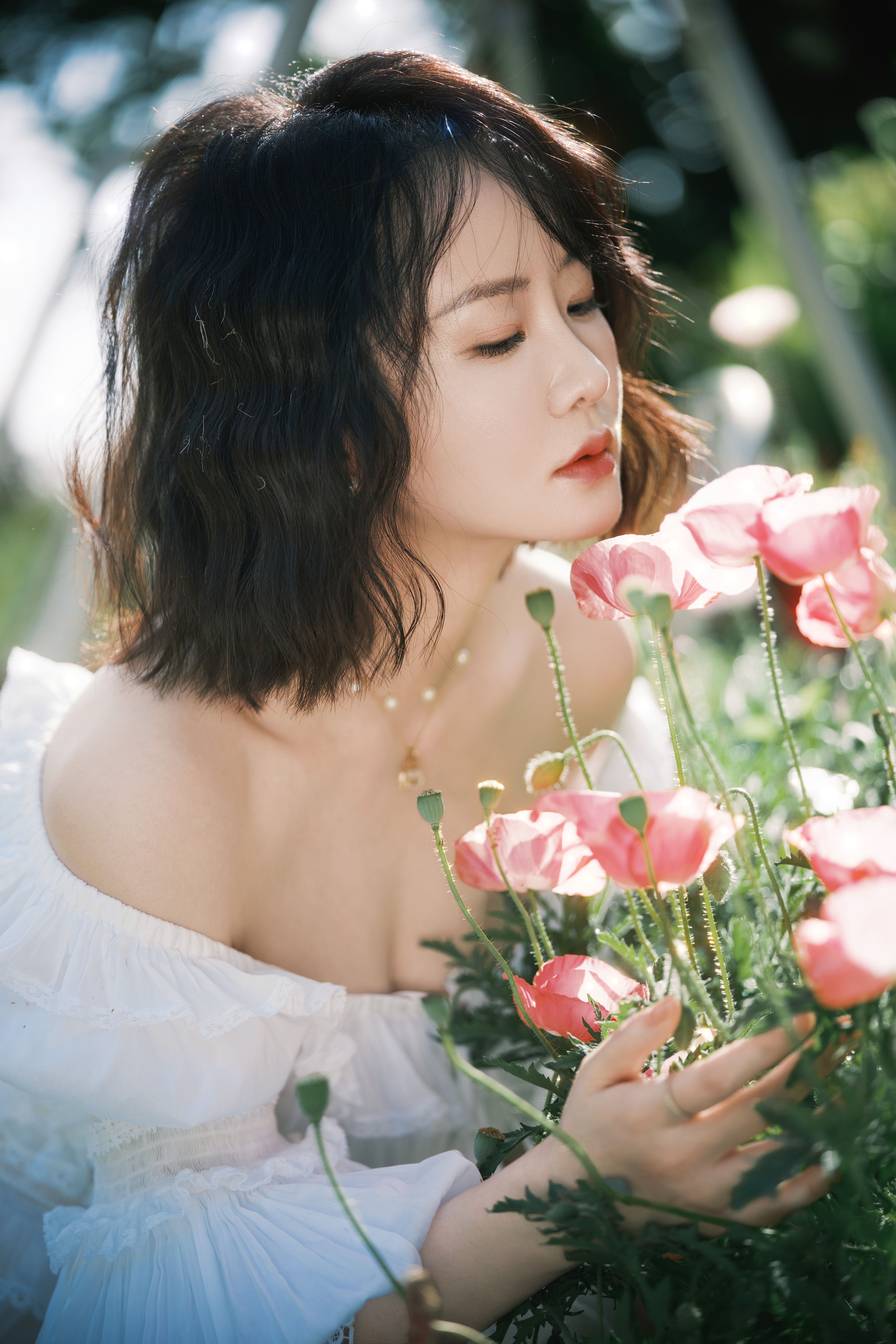 A young woman in a white dress admires vibrant pink flowers outdoors.