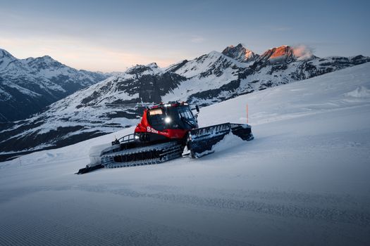 A snow groomer prepares ski slopes against the backdrop of the Swiss Alps during winter dusk.