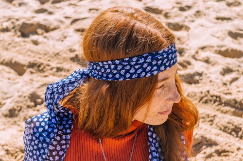 Photo of Woman With Freckles in Blue Floral Headscarf and Orange Top Looking Away While Sitting on Sand