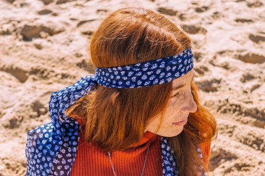 一位戴着花卉头巾的红发女人体贴地坐在沙滩上，散发着夏日的气息