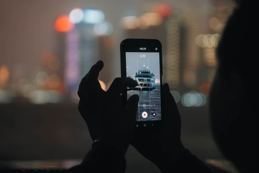 Silhouette holding smartphone capturing distant cityscape at night, bokeh lights in background.